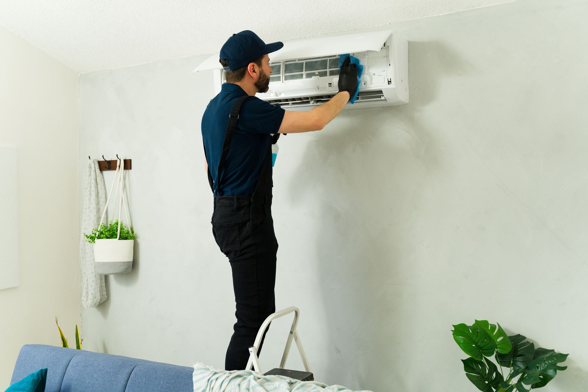 Man in uniform cleaning an AC unit on a wall, standing on a step stool.