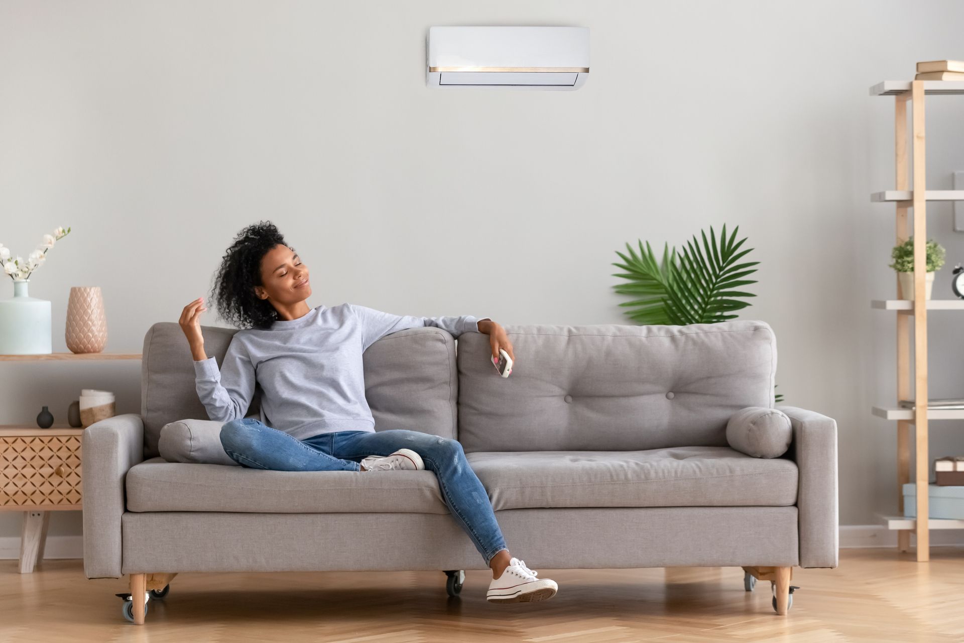 Woman relaxing on a couch, using AC remote. Light gray interior with a shelf.