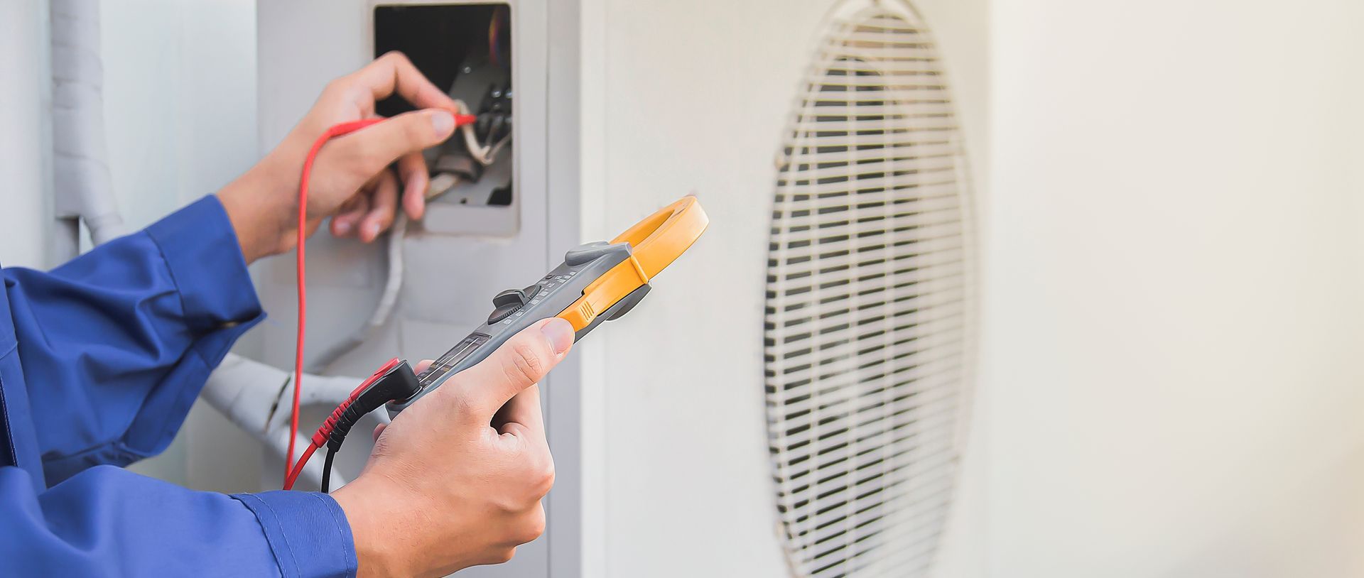 An electrician in a blue jumpsuit tests electrical wiring on an air conditioning unit.