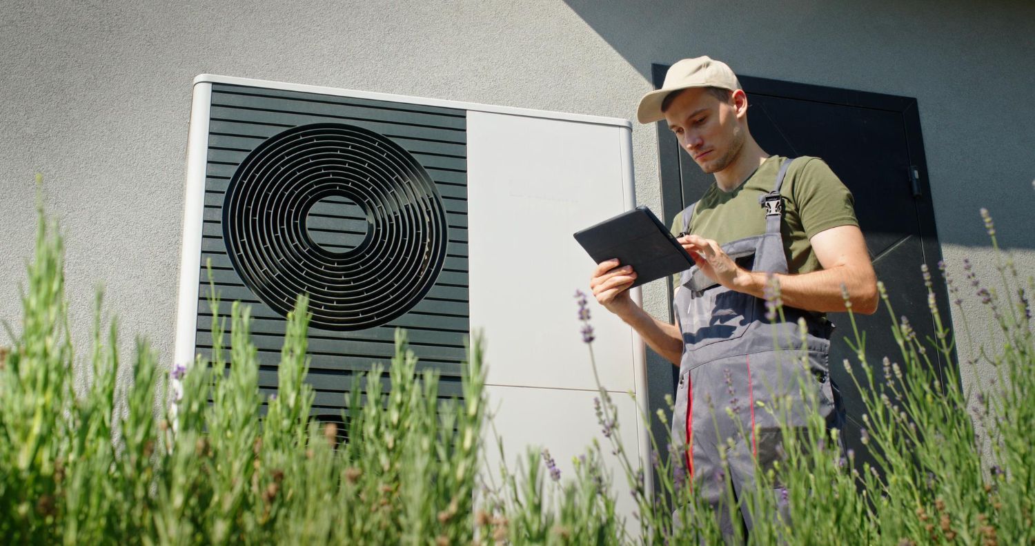 A technician in overalls and hat uses a tablet next to an outdoor HVAC unit.