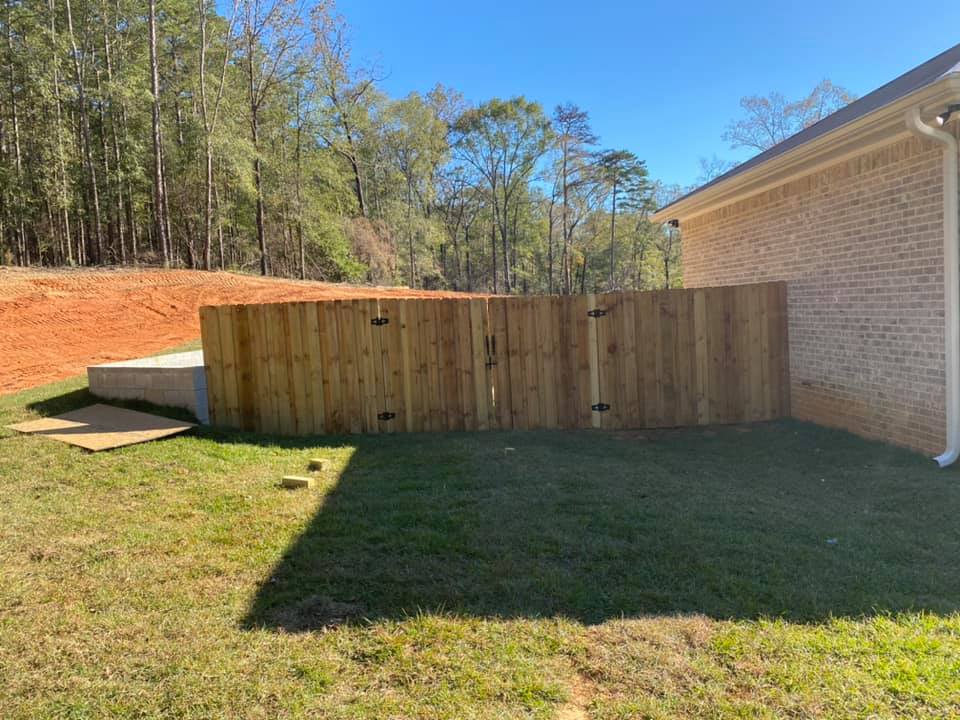 Wooden fence surrounds a backyard, next to a brick house and grassy area. Sunny, outdoor setting.