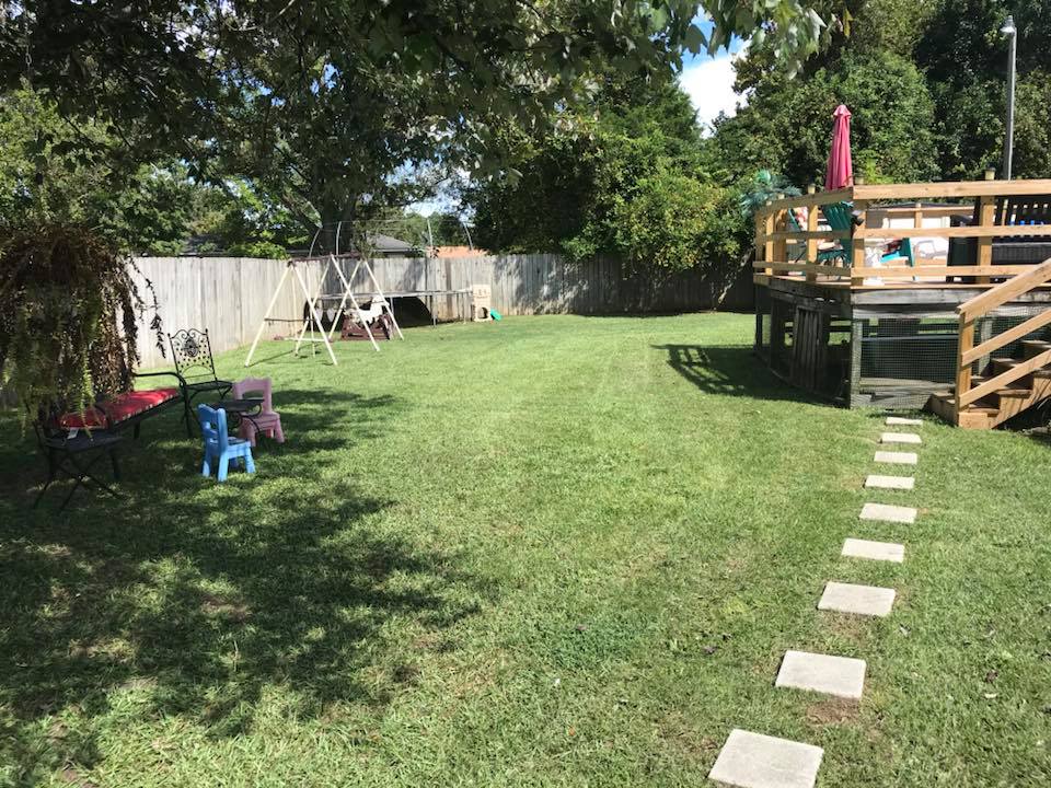 Grassy backyard with a wooden deck, stepping stones, trees, fence, and small child chairs.