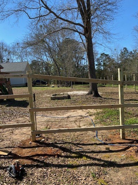 Wooden three-rail fence in a yard, gate open. Sunlit trees and house in the background.