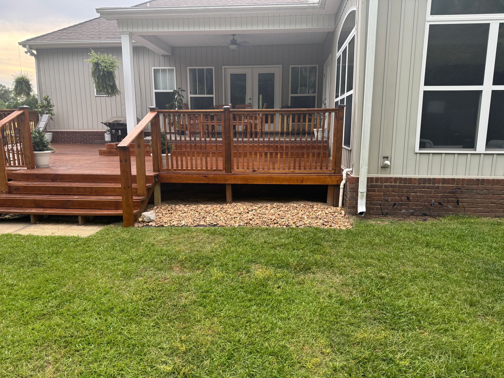 Wooden deck with brown stain, overlooking green lawn and beige gravel. House in background.