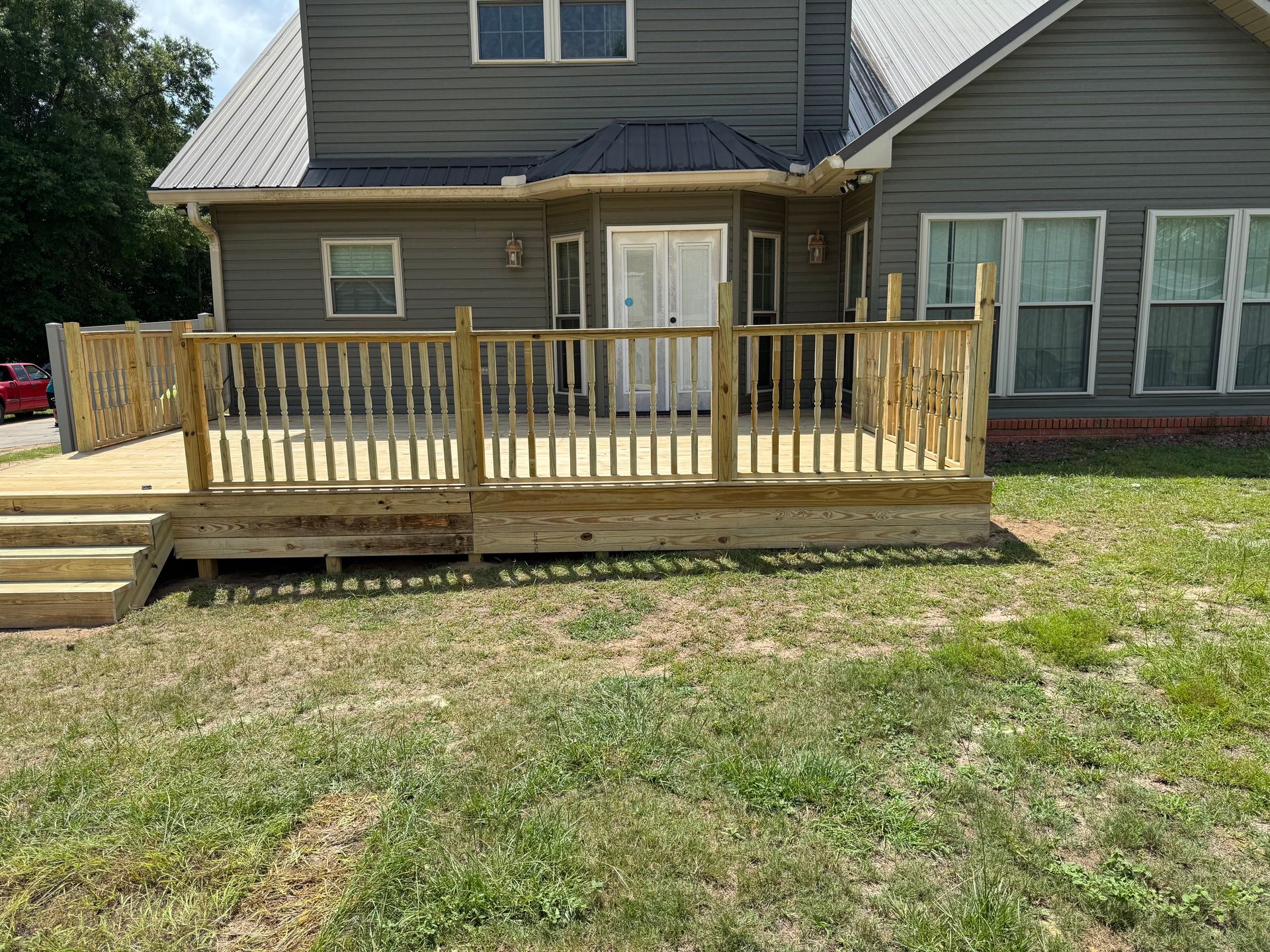Wooden deck attached to a gray house with railings and stairs, surrounded by grass.