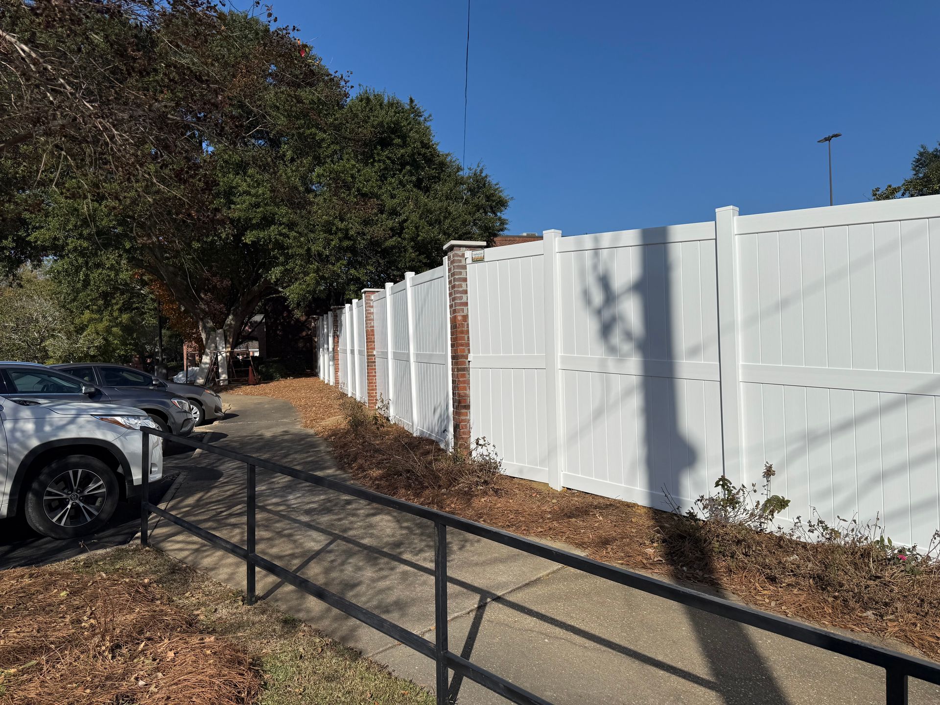 White fence with brick pillars along a pathway; cars parked on the left.