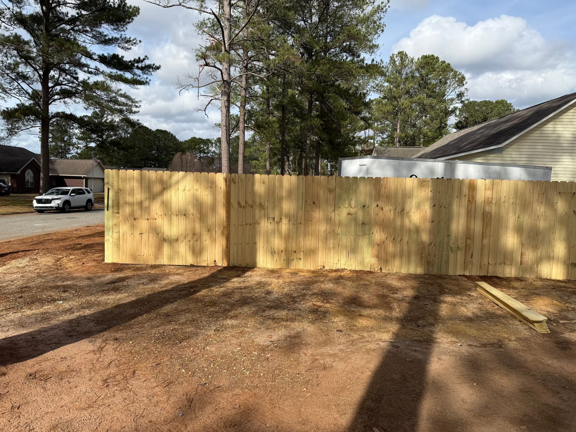 New wooden fence in front of a house, with a dirt yard and trees in the background.