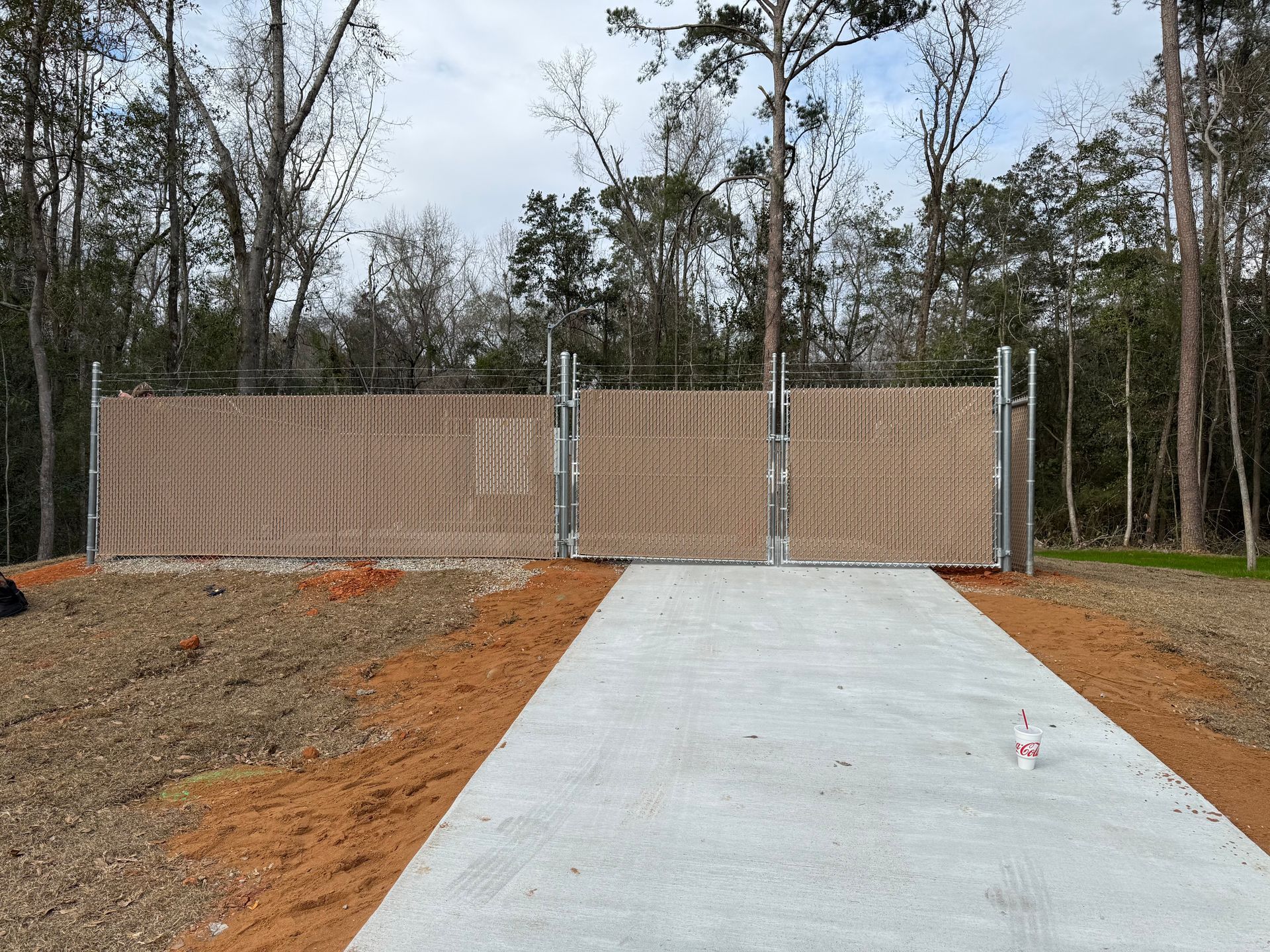 Concrete driveway leading to closed wooden gates, framed by metal posts; wooded background.