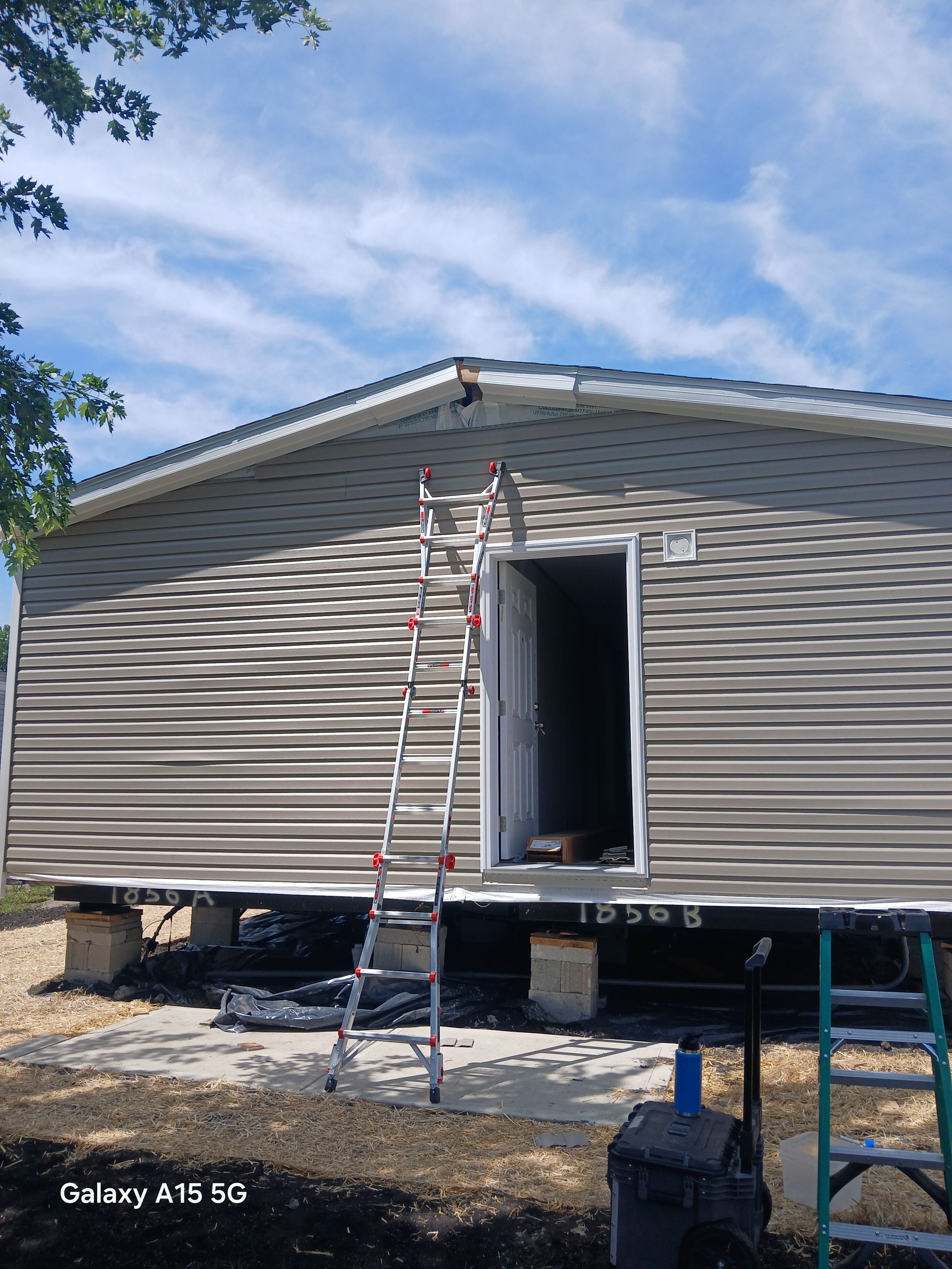 Ladder leaning against house with open door, gray siding, blue sky.