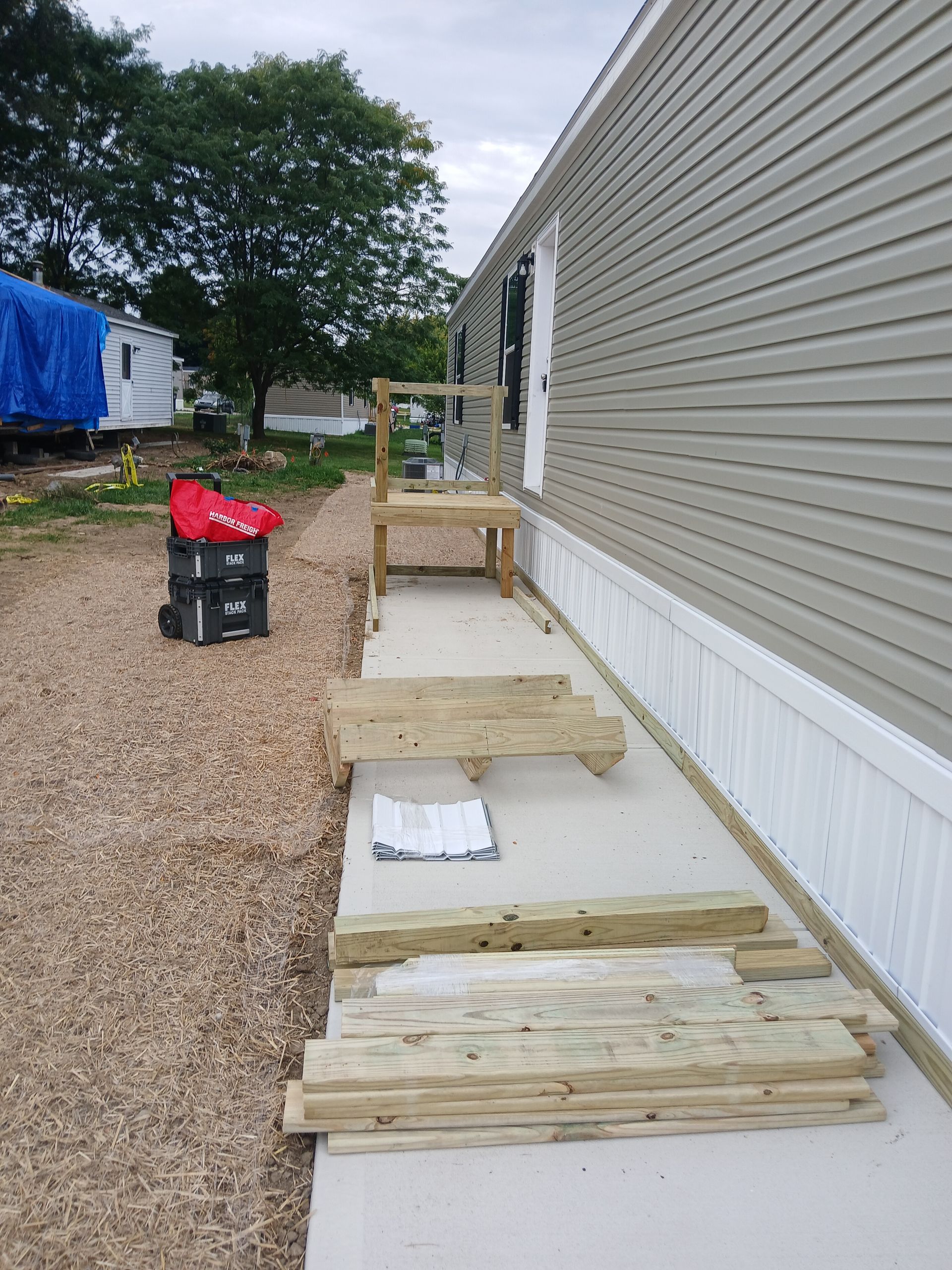 Ramp construction at a mobile home entrance: wood materials, concrete path, gray siding, and gravel ground.