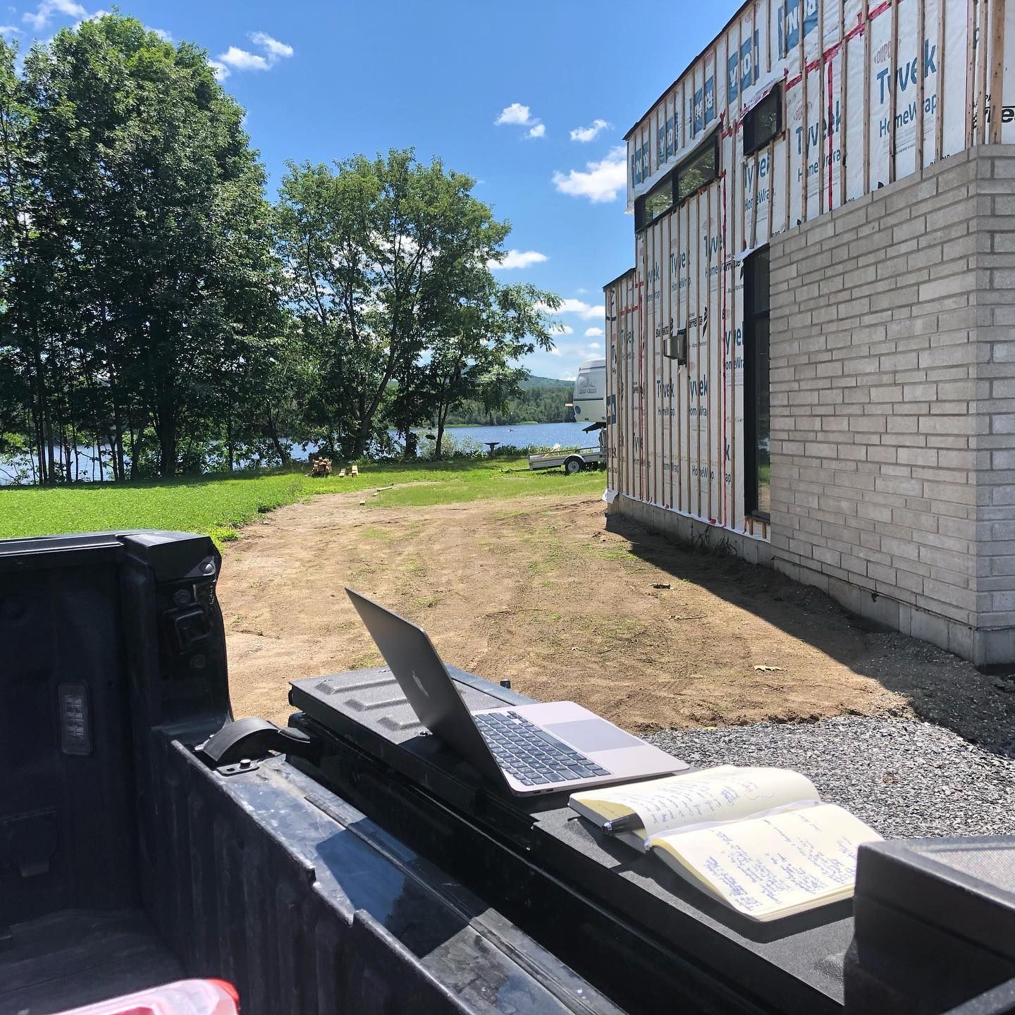 Un ordinateur portable et un cahier ouvert sont posés sur le hayon d'un camion, donnant sur un chantier ensoleillé près d'un lac.