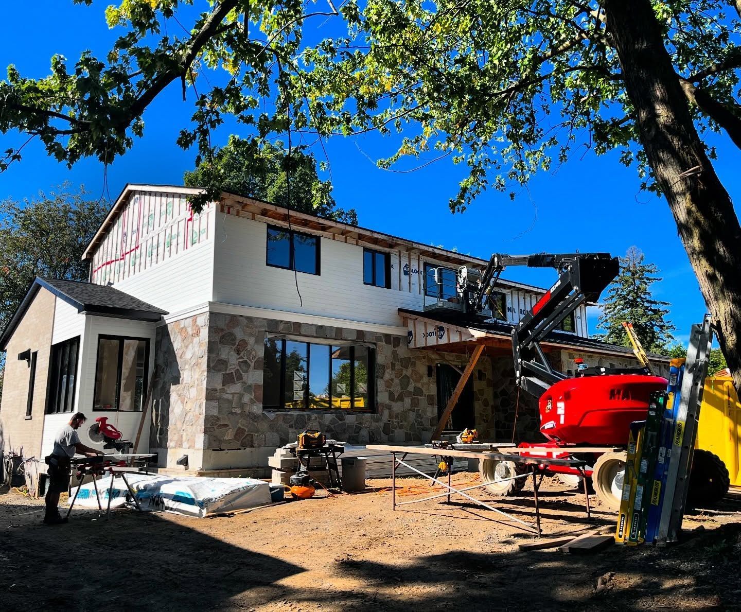 Un ouvrier coupe du bois sur un chantier de construction, une nacelle élévatrice rouge étant garée devant une maison en rénovation.