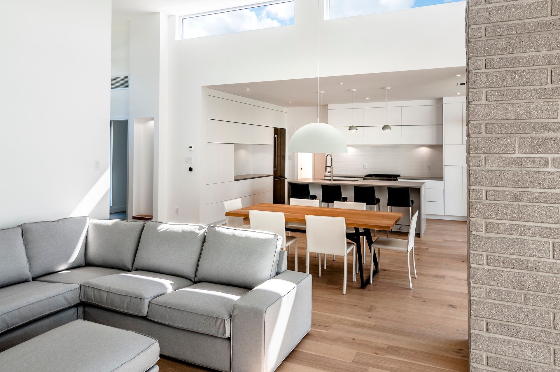 Espace de vie moderne et ouvert avec canapé d'angle gris, table à manger et cuisine blanche. Parquet et briques apparentes.