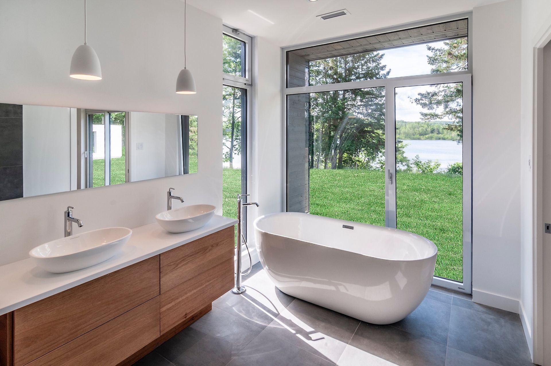 Salle de bains moderne avec baignoire blanche, meuble-lavabo en bois et grande fenêtre donnant sur la verdure et l'eau.