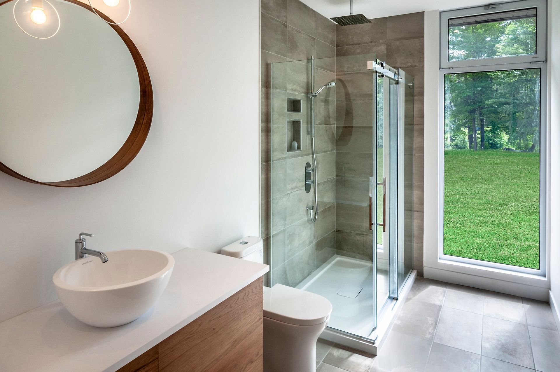 Salle de bains moderne avec douche, lavabo, toilettes, grand miroir rond et fenêtre donnant sur la verdure.