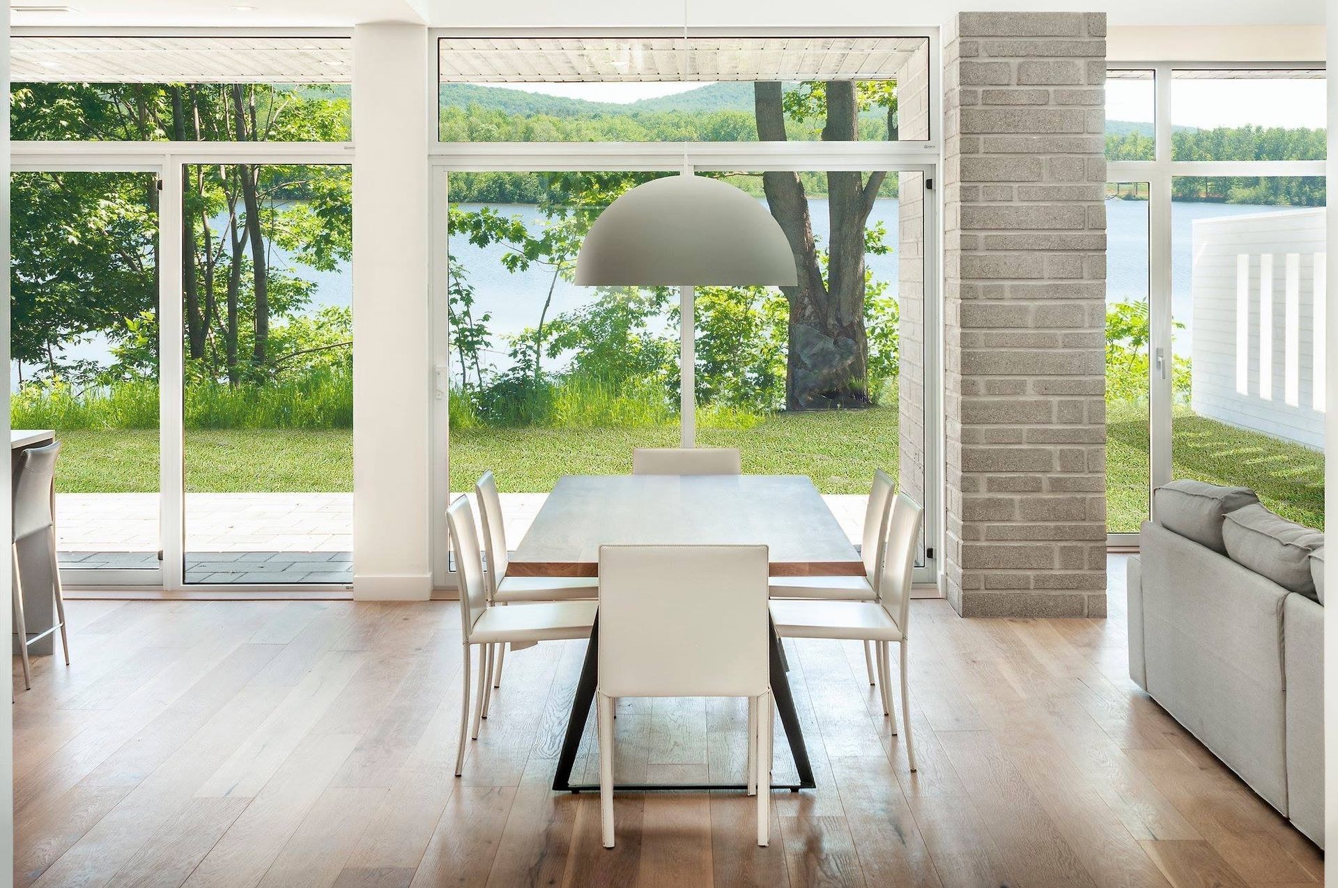 Salle à manger avec de grandes fenêtres donnant sur un lac et des arbres. Table et chaises avec une suspension lumineuse.