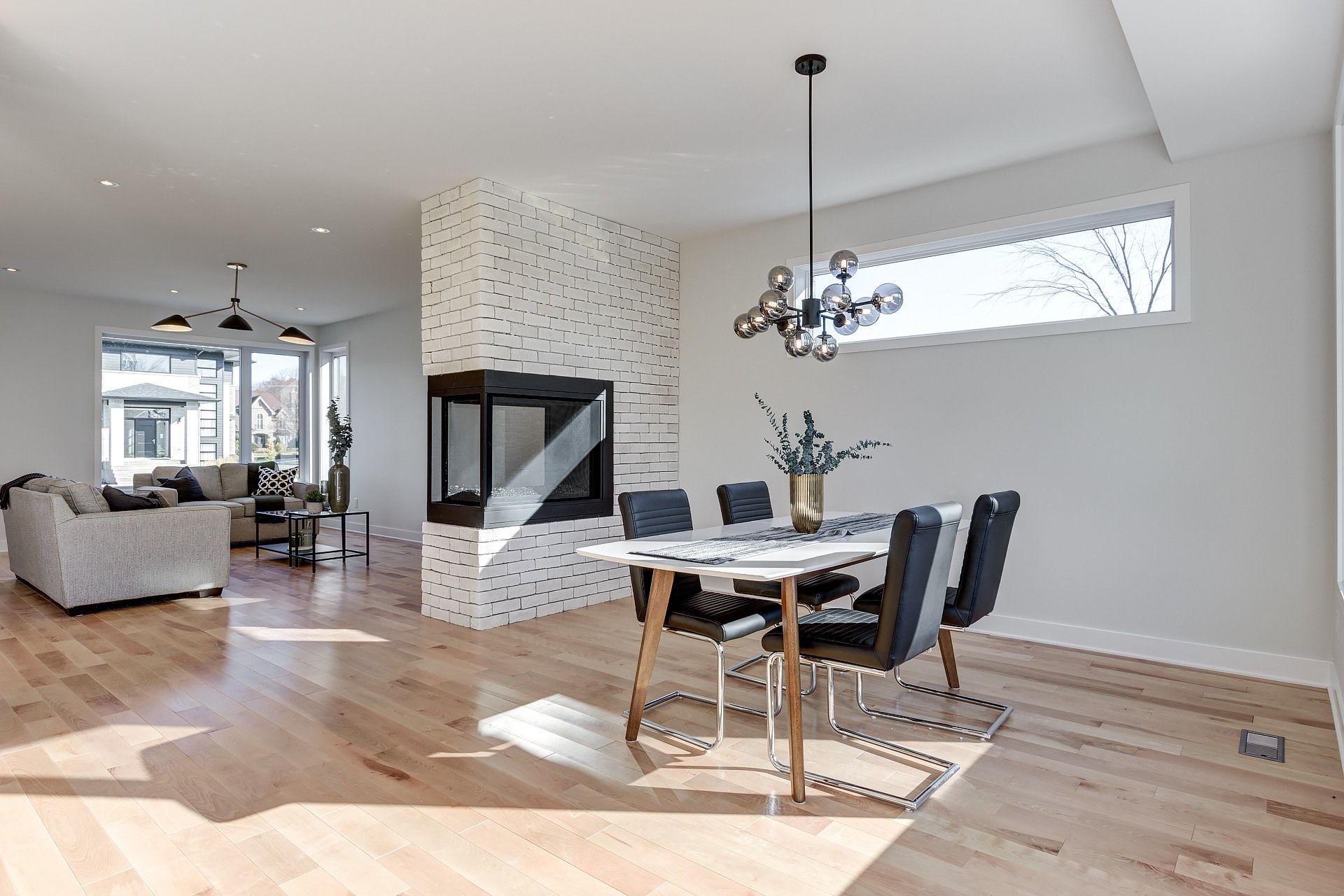 Salle à manger moderne avec table, chaises et lustre ; cheminée et salon visibles.