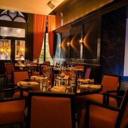 Intérieur de restaurant avec tables rondes, chaises et rideaux orange, murs sombres et fenêtres.