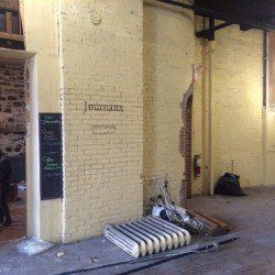 Intérieur en briques jaunes avec inscription « Journaux ». Tableau noir, radiateur et débris au sol.