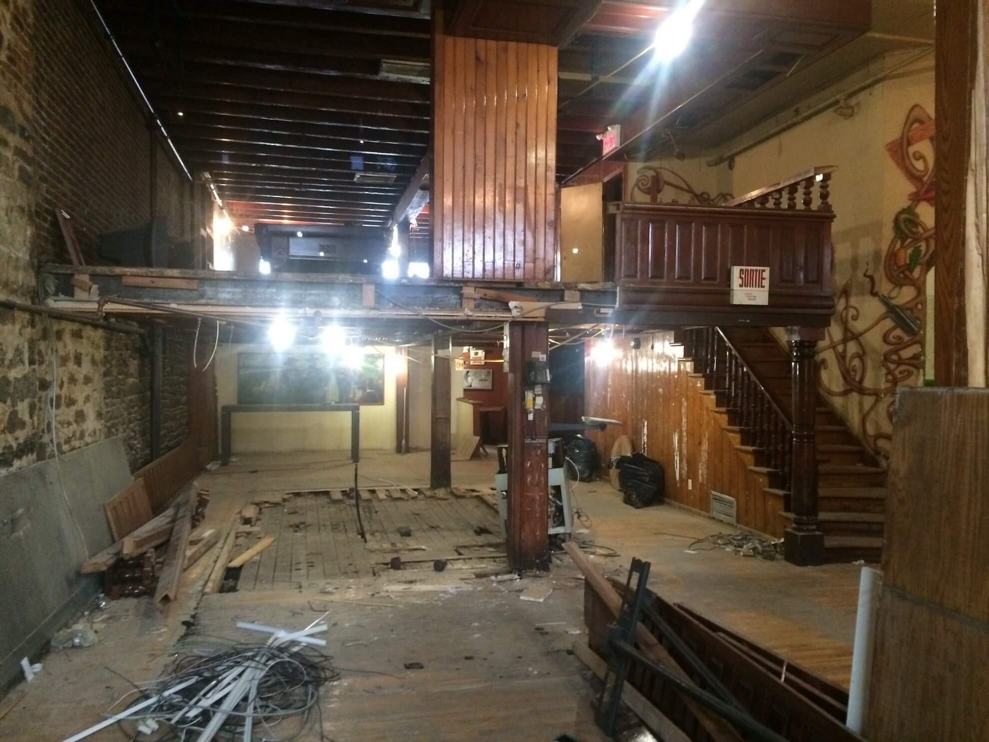 Vue intérieure d'une pièce en rénovation avec une terrasse en bois surélevée, un escalier, des murs en briques apparentes et des débris.