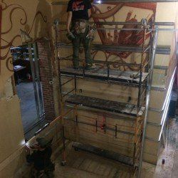 Une personne sur un échafaudage peint un mur. Une autre personne en dessous observe. Décoration intérieure avec œuvres d'art murales.