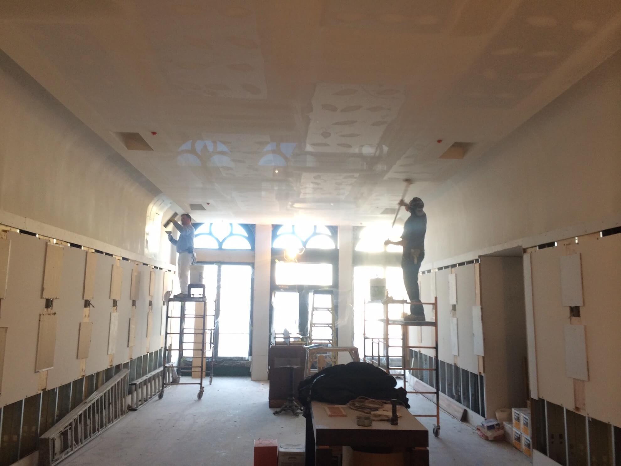 Deux ouvriers installent des cloisons sèches dans une pièce avec de grandes fenêtres, des échelles et des montants exposés.