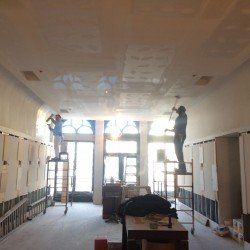 Deux ouvriers plâtrant une grande pièce avec de hauts plafonds et de grandes fenêtres.