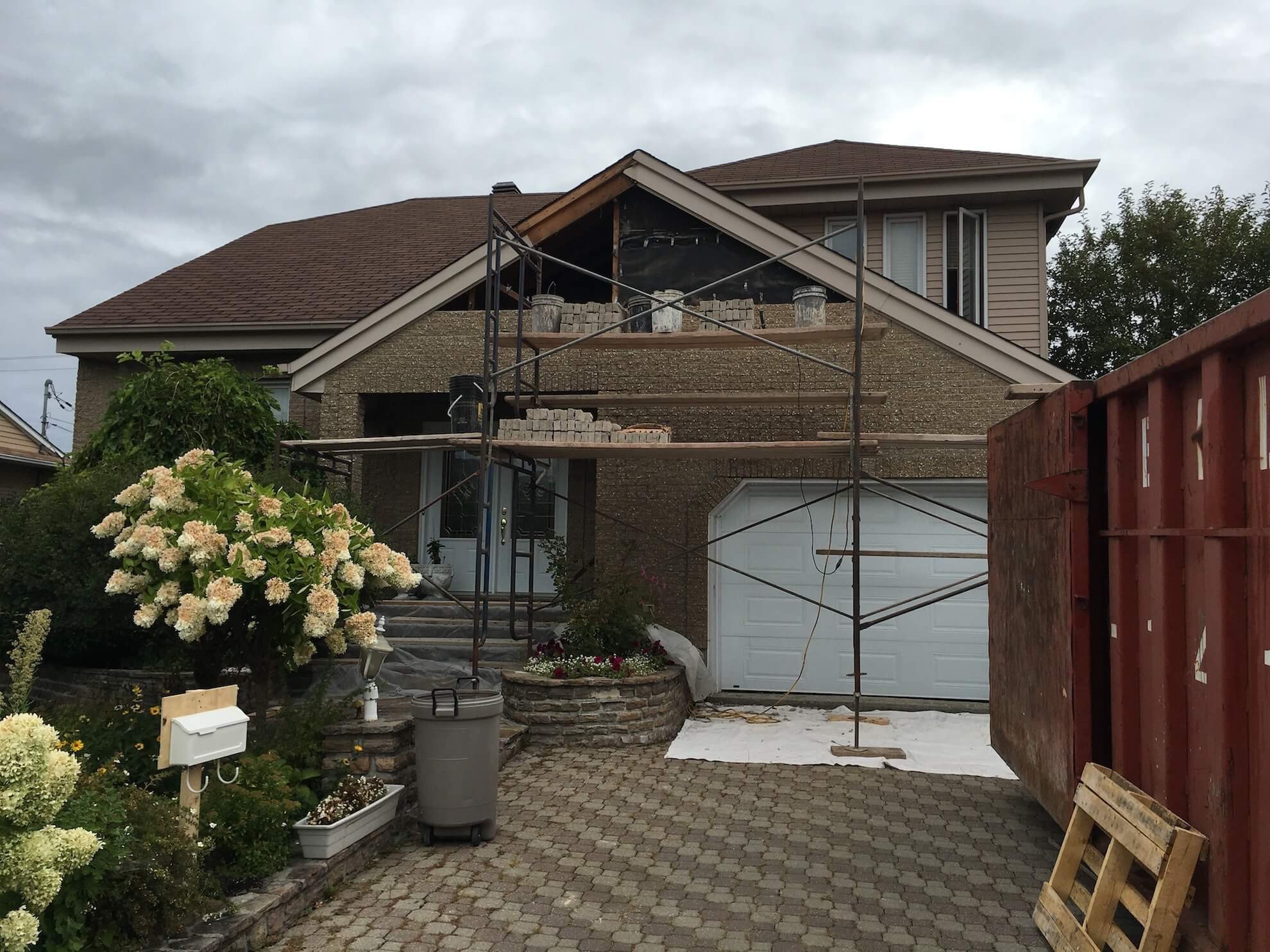 Une maison d'habitation en construction, avec des échafaudages dans l'allée d'entrée et une grande benne à ordures rouge sur le côté.