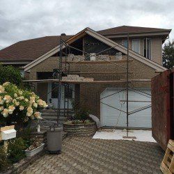 Extérieur de la maison en construction, échafaudage devant. Maçonnerie en cours d'ajout. Toiture brune, journée nuageuse.