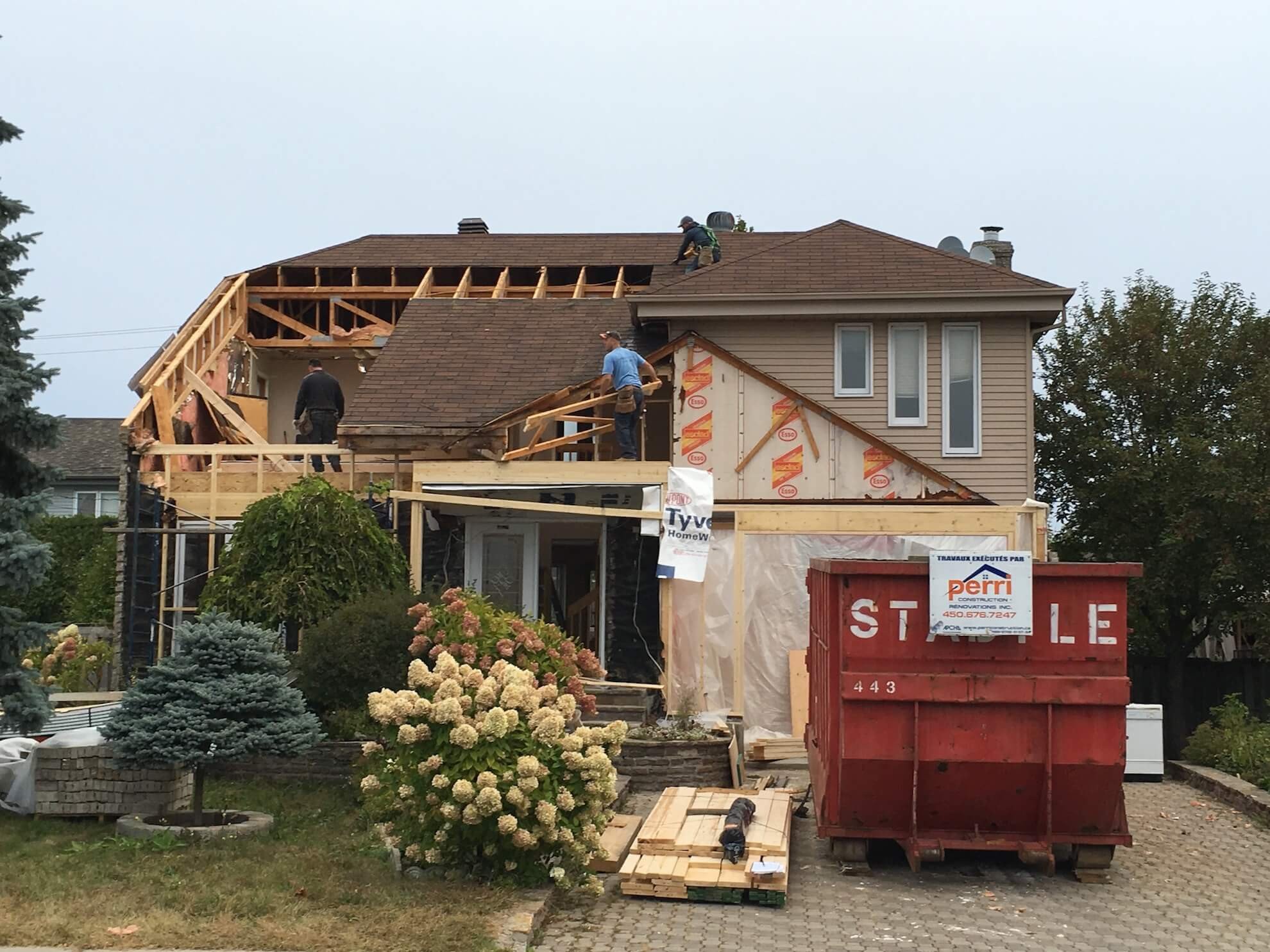 Des ouvriers effectuent des travaux de rénovation extérieure sur une maison d'habitation de deux étages avec un toit ouvert et une grande poubelle rouge.