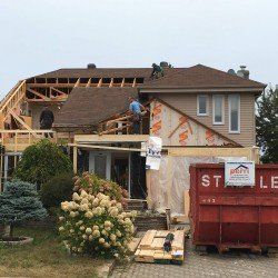 Construction d'une maison à deux étages; ouvriers sur le toit et l'échafaudage; benne à ordures rouge dans la cour avant.