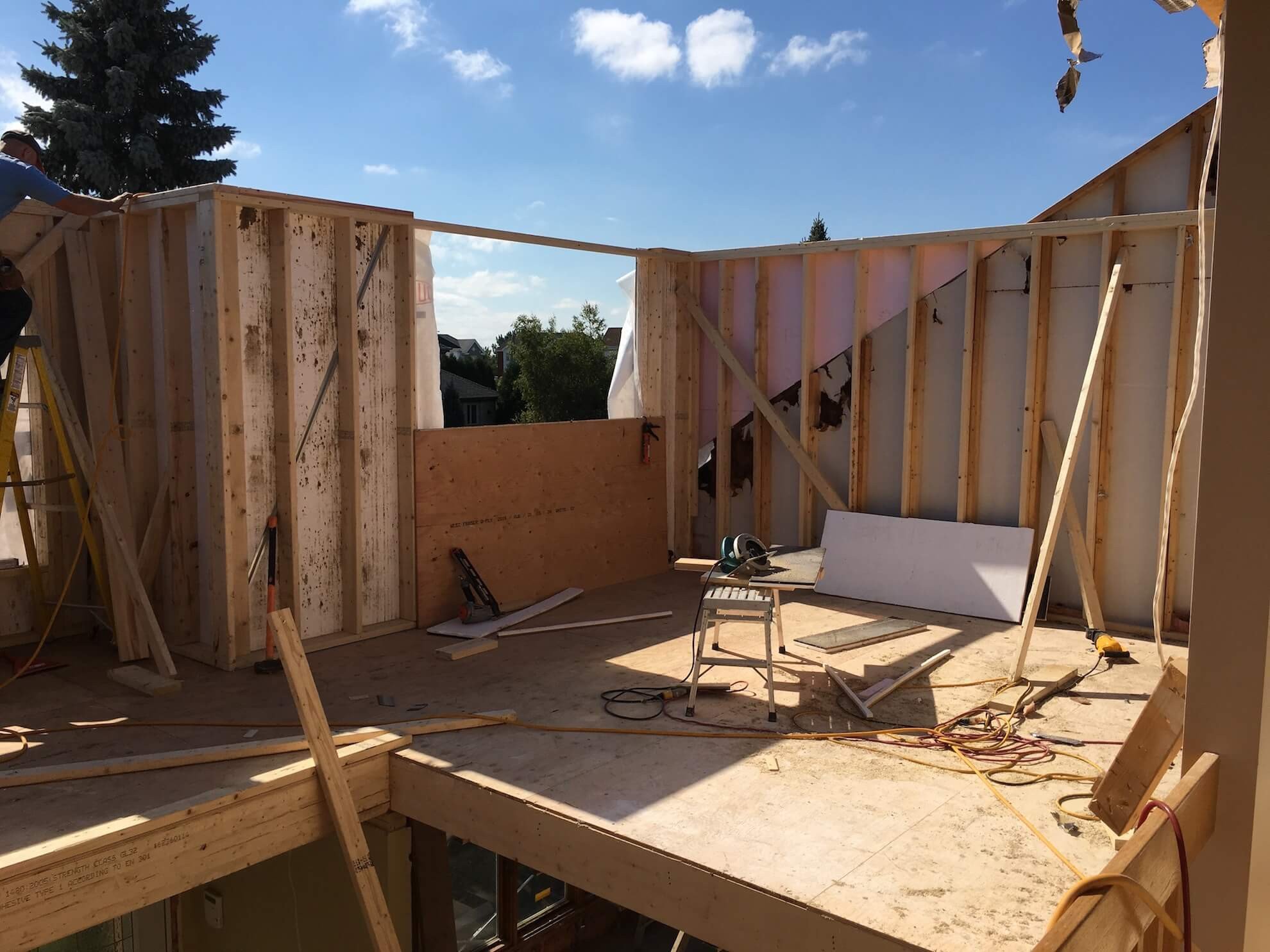 La charpente en bois inachevée d'une extension de bâtiment en construction, avec ses montants et panneaux de contreplaqué apparents, sous un ciel bleu.