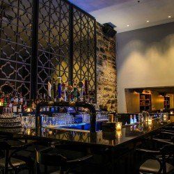 Un bar faiblement éclairé avec un comptoir noir, des panneaux métalliques décoratifs et des bouteilles d'alcool.