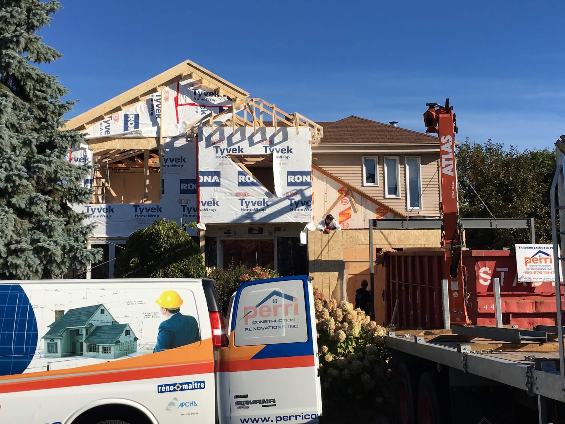 Un chantier de construction avec une maison à deux étages partiellement construite, un camion de service stationné et une grue orange se détachant sur un ciel bleu.
