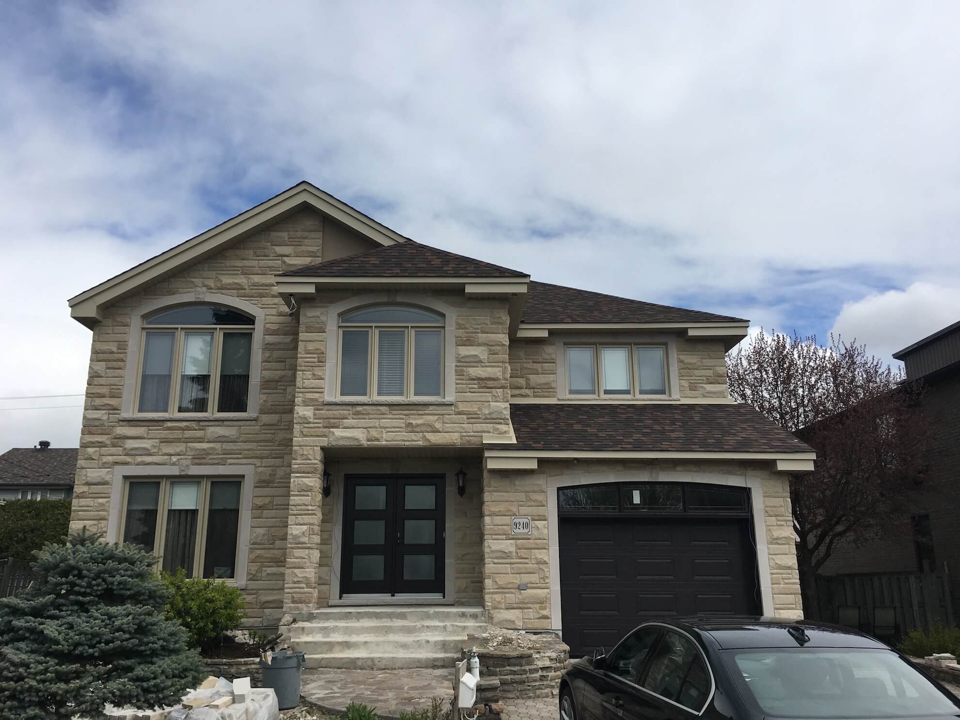 Maison en pierre de deux étages avec toit et porte de garage brun foncé ; voiture noire garée devant.