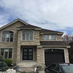 Maison à deux étages avec façade en pierre, porte noire, toit foncé et garage attenant; voiture garée dans l'allée.