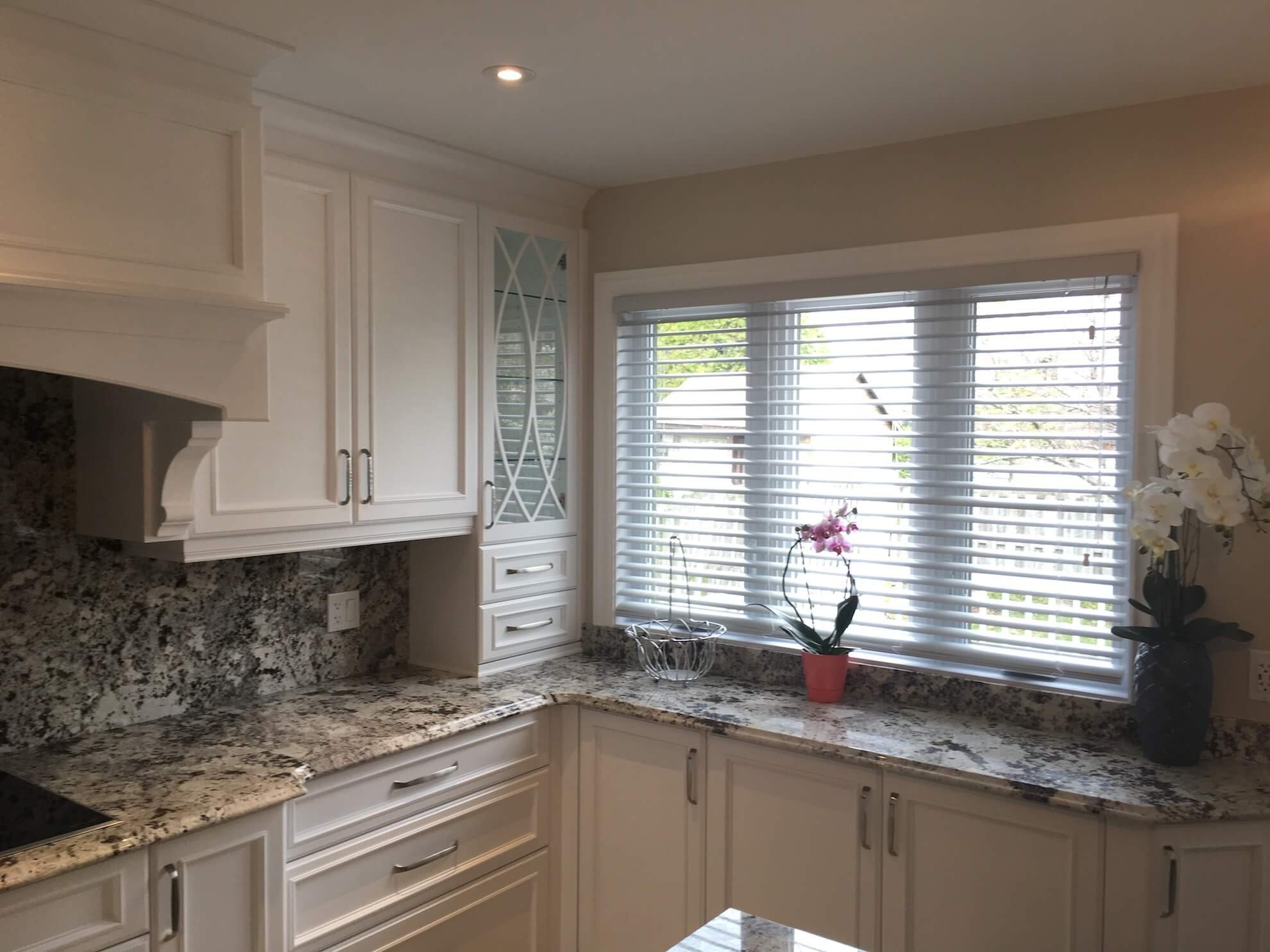 Une cuisine lumineuse avec des armoires blanches, des comptoirs en granit, une fenêtre avec des stores gris et une orchidée en pot.