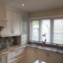 Cuisine blanche avec comptoirs en granit, armoires blanches et fenêtre avec stores.