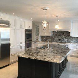 Cuisine blanche avec îlot foncé et comptoir en granit, électroménagers en acier inoxydable et deux luminaires suspendus.