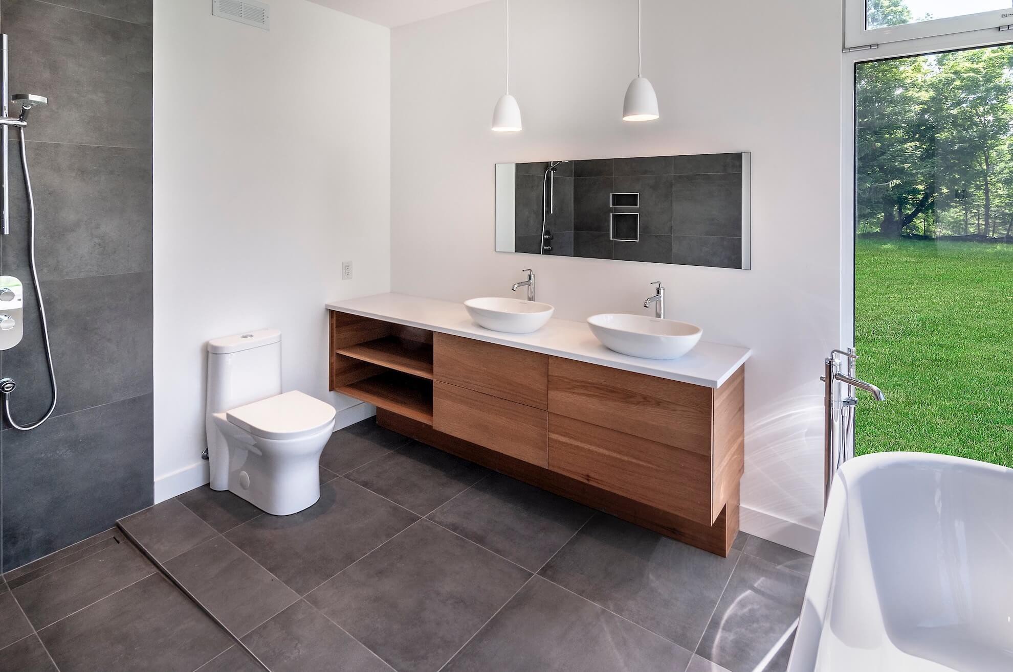 Salle de bain moderne avec vanité en bois, double vasque, carrelage gris et grande fenêtre donnant sur la pelouse verte.