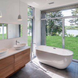 Salle de bain moderne avec vanité en bois, lavabos blancs et baignoire face à une grande fenêtre donnant sur une pelouse verte et de l'eau.