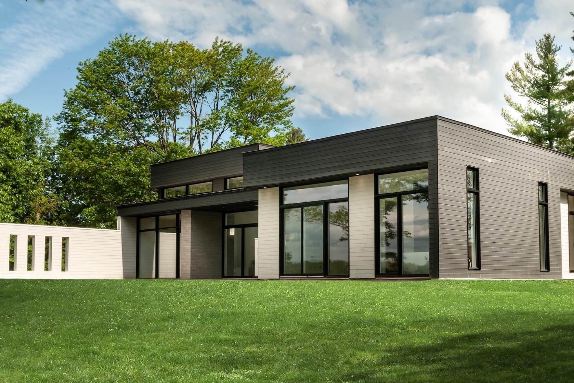 Maison moderne avec toit noir, grandes fenêtres, revêtement gris et pelouse verte.