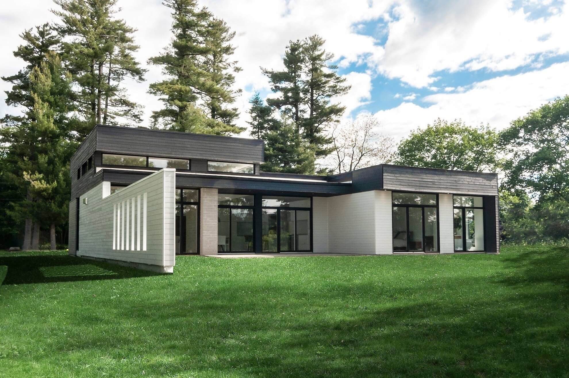 Maison moderne avec des murs blancs, des garnitures noires et de grandes fenêtres sur une pelouse verte sous un ciel bleu.