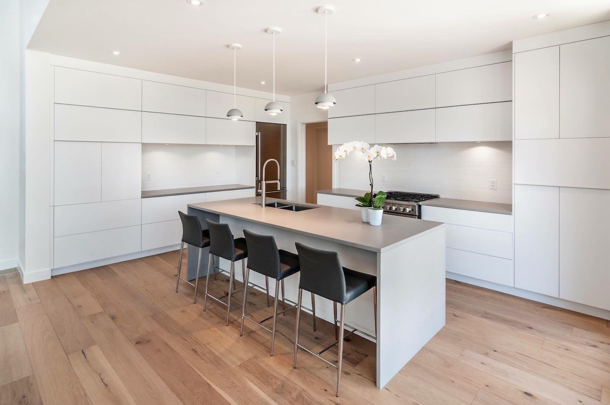 Cuisine blanche moderne avec îlot, tabourets, planchers de bois franc et éclairage au plafond.