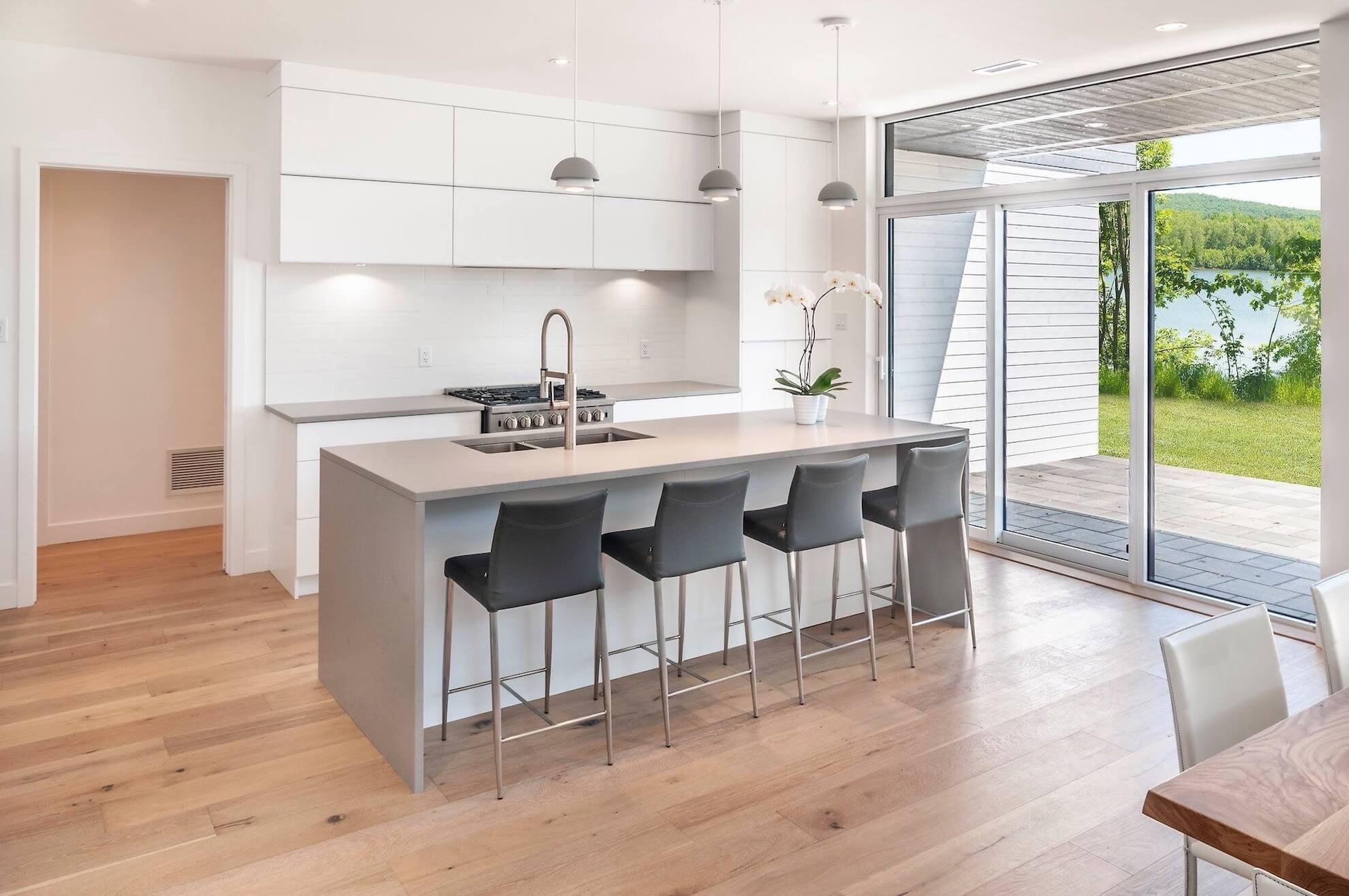 Cuisine blanche moderne avec îlot, tabourets gris et grande fenêtre donnant sur le lac.
