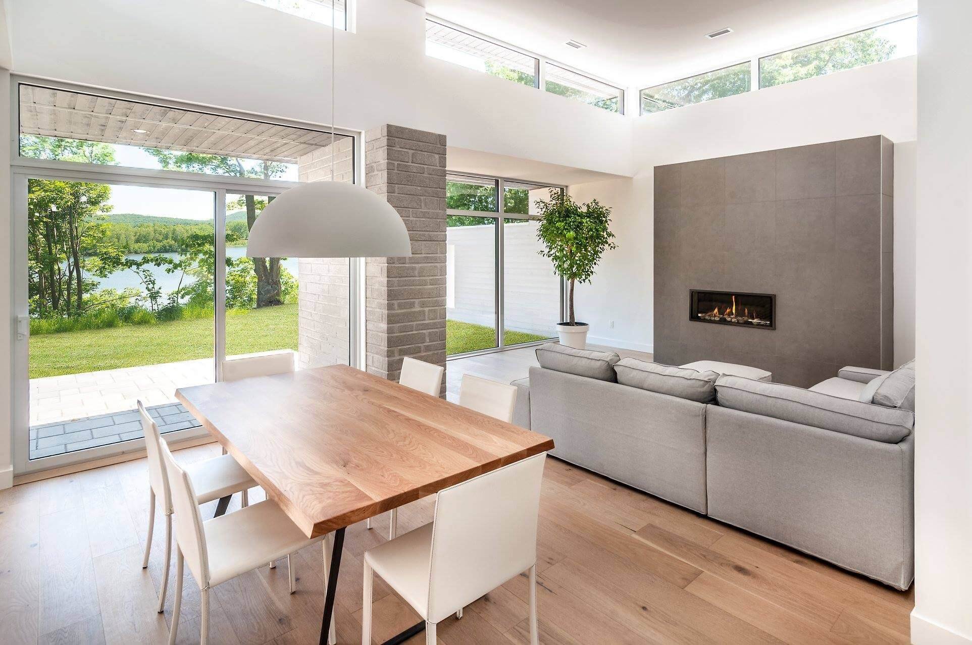 Salle à manger/salon moderne avec vue sur le lac ; table en bois, canapé gris, cheminée et murs blancs.