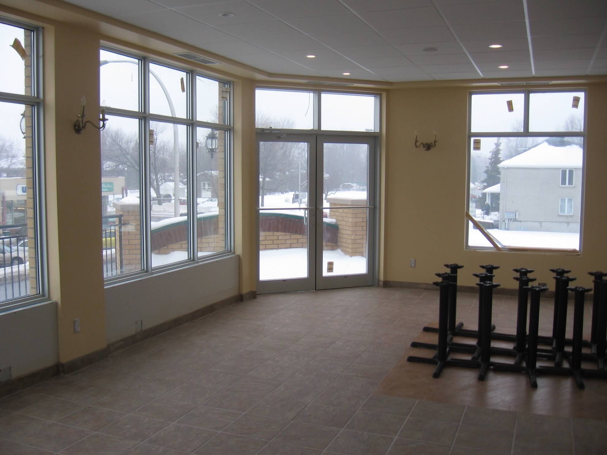 Pièce vide et ensoleillée, avec de grandes fenêtres donnant sur une rue enneigée. Pieds de table empilés à droite.