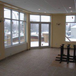 Pièce vide avec de grandes fenêtres donnant sur une rue enneigée. Des pieds de table noirs sont posés au sol.