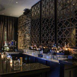 Bar avec comptoirs noirs, panneaux muraux en miroir, bouteilles d'alcool et verres ; intérieur sombre et élégant.