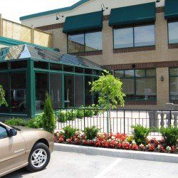 Voiture beige garée devant un bâtiment avec des auvents verts, une véranda en verre et un parterre de fleurs.